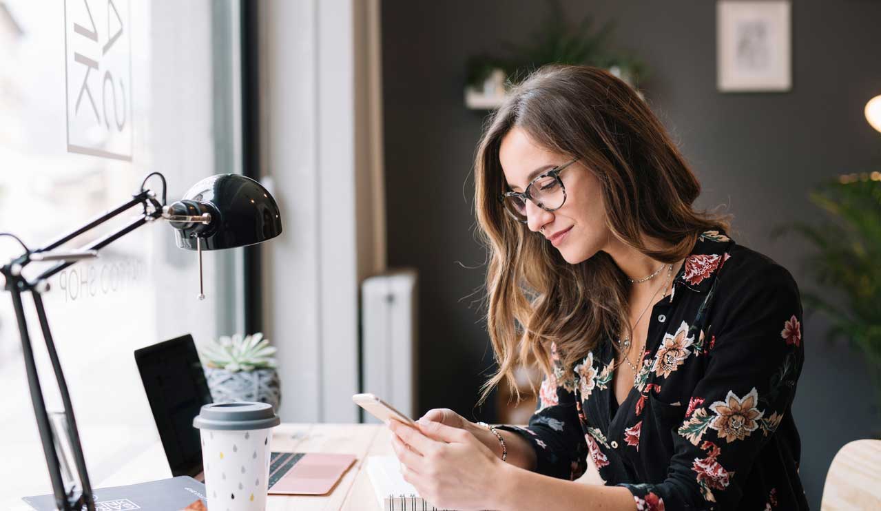 Woman with glasses at her desk looks at her mobile phone with a laptop in front of her and a large desk lamp and coffee cup