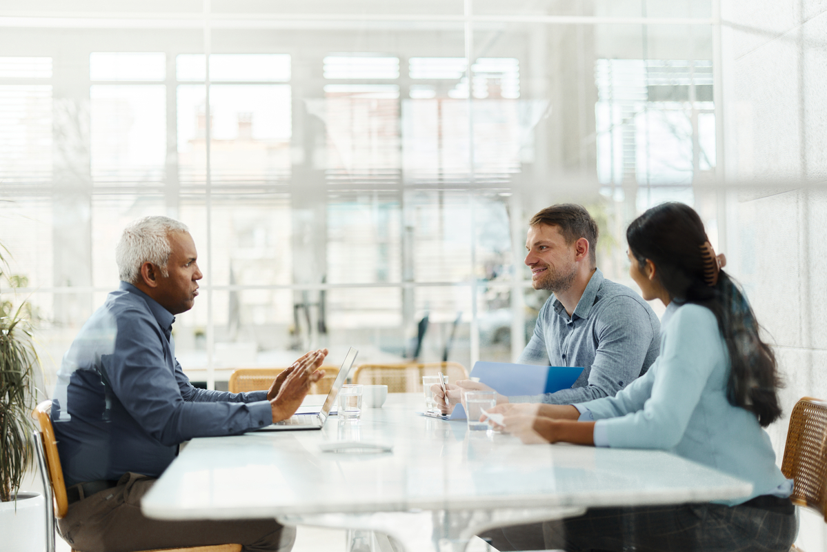 In a modern conference room, a senior Southeast Asian man looks thoughtfully across the table at a smiling white and Southeast Asian woman, who appear to be junior associates