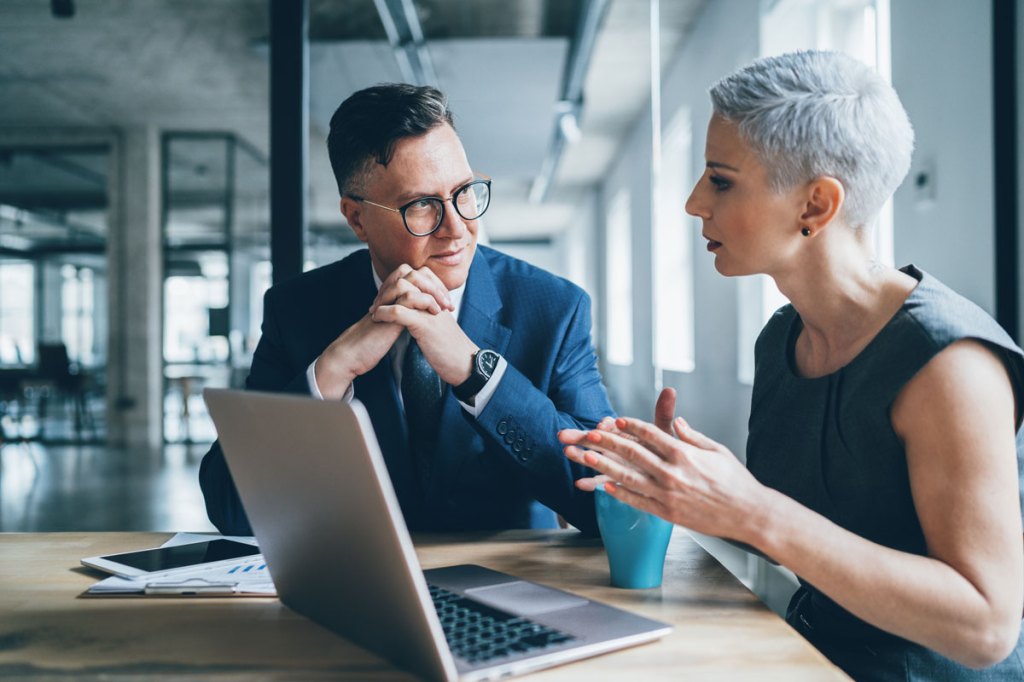 Business man and woman working together at a laptop on a conference table in an office. Man has glasses, suit and watch, woman has white hair and sleeveless top with a blue mug underneath her. Man has a tablet and clipboard on the table near him