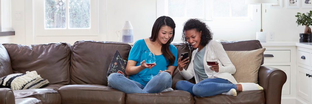 Two females on couch holding wine glass & looking at cell phone