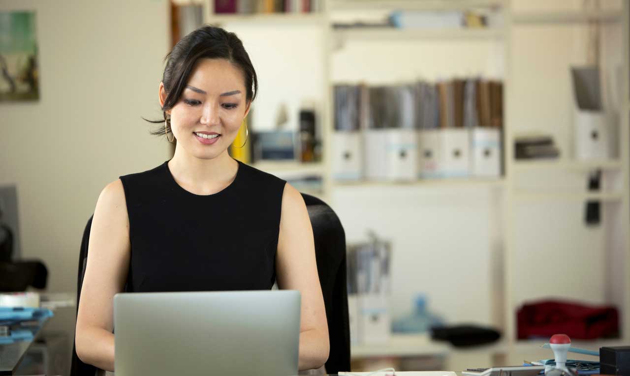 An Asian woman with dark hair and black tank top looking at her laptop