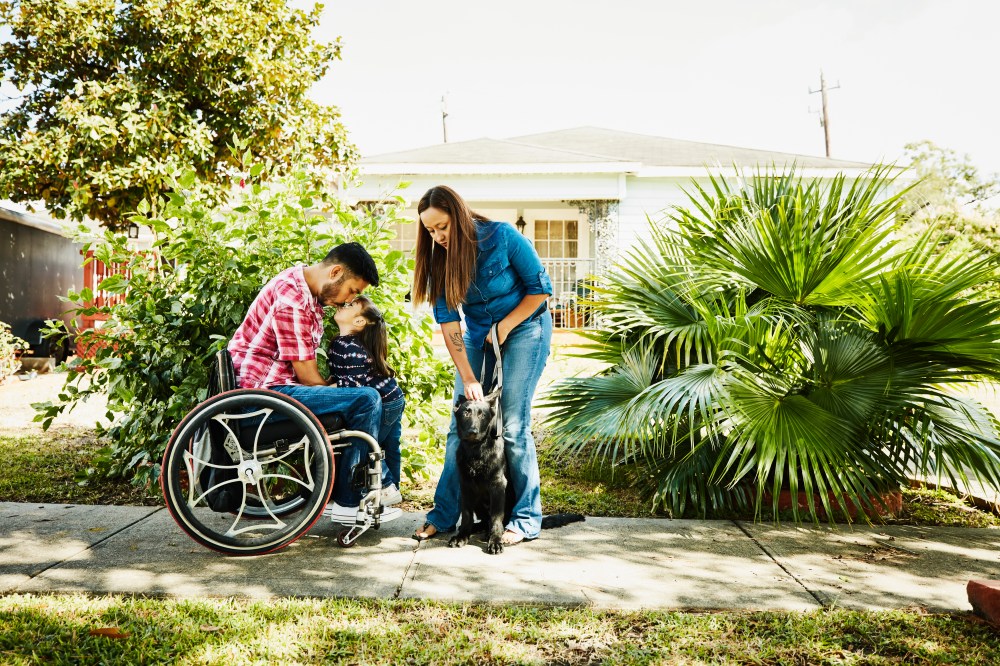 Young daughter giving father in wheelchair a kiss while standing in front of home with mother and dog