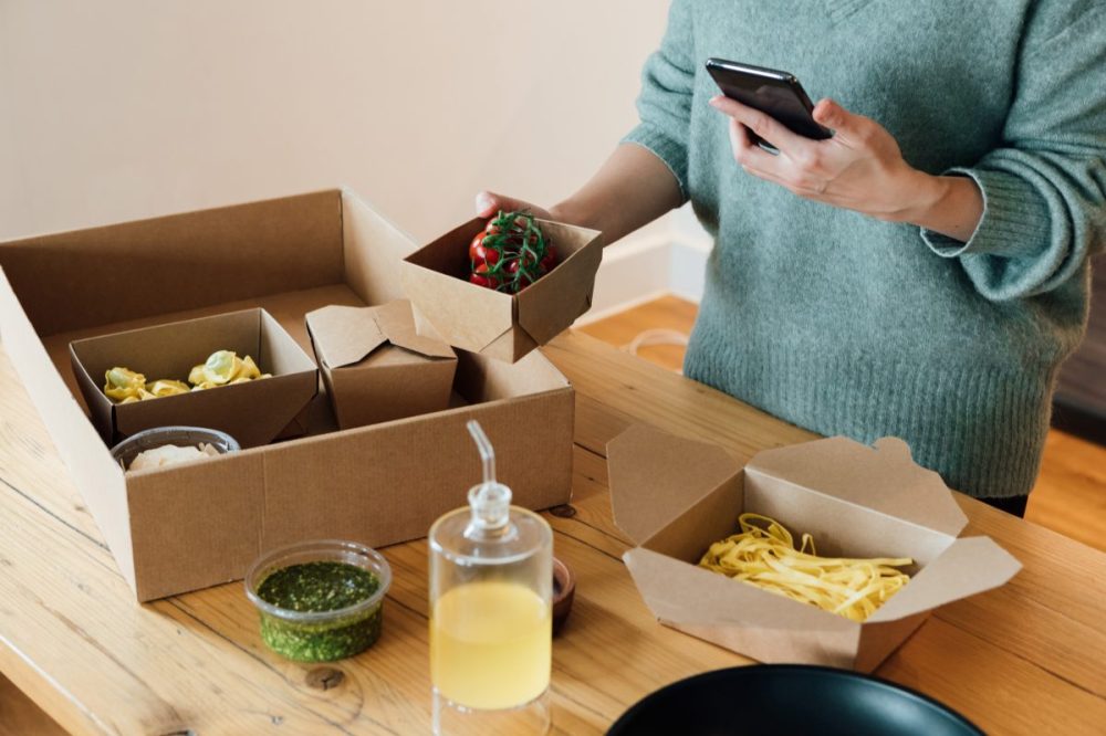 A woman holding a phone stands by a counter with small to-go boxes on top of it.