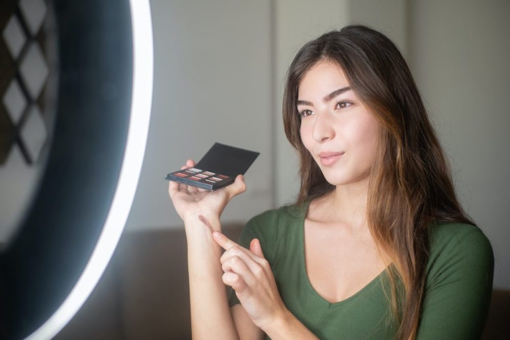 A woman applies face makeup with a brush while standing in front of a bright ring light.