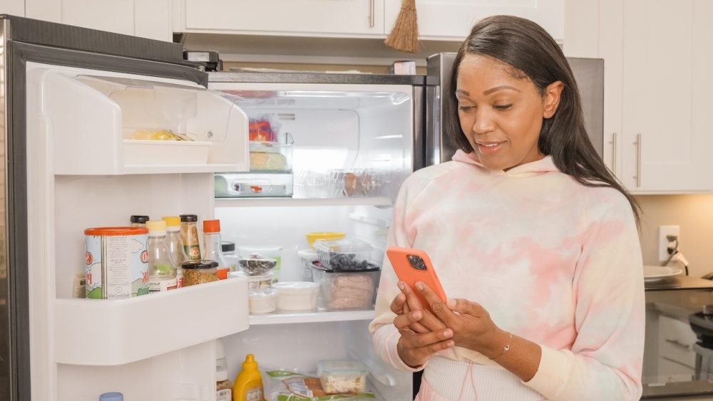A black woman stands in front of her refrigerator online shopping from her phone