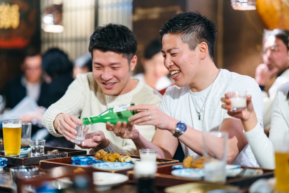 Two young Asian American consumers pour a drink white sitting at a table, talking and laughing.