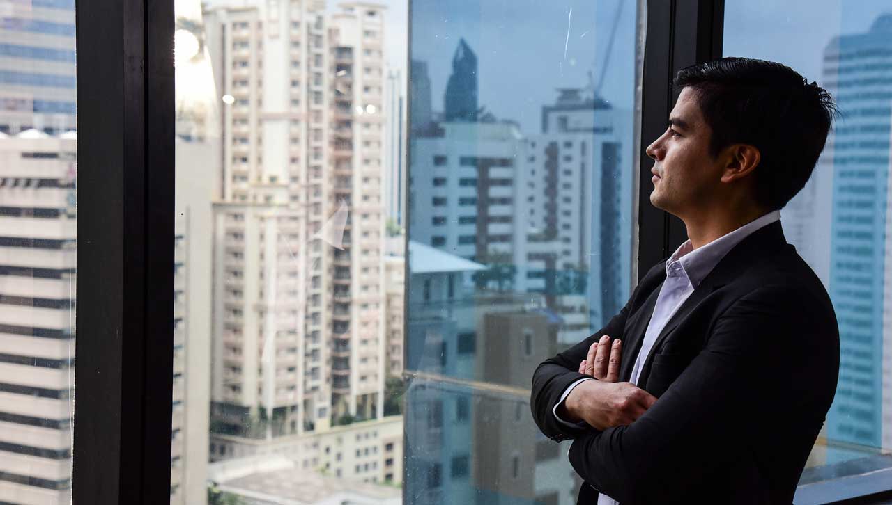 Man in suit with dark hair looking out an office window at a city with tall buildings.
