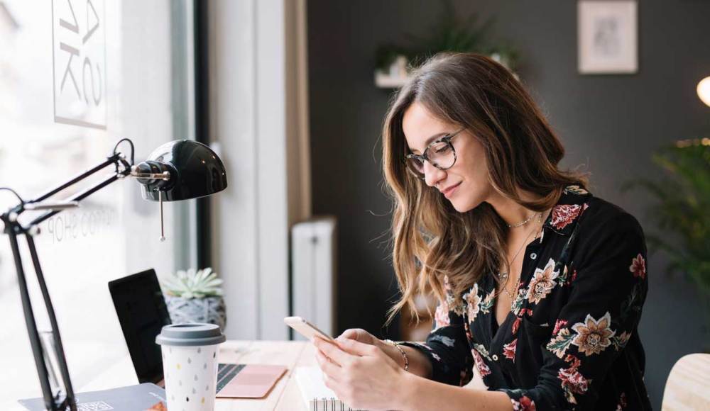 Woman with glasses at her desk looks at her mobile phone with a laptop in front of her and a large desk lamp and coffee cup