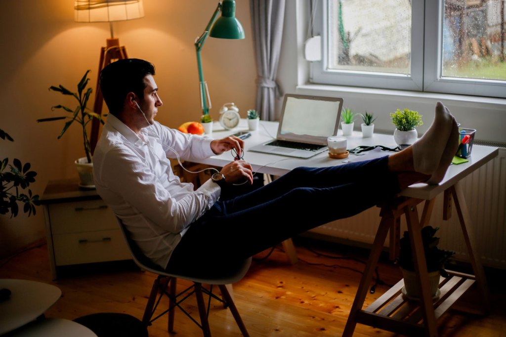 Man with white shirt, blue jeans, and white socks holding pair of glasses with earbuds looking at laptop on desk with a green lamp overlooking window
