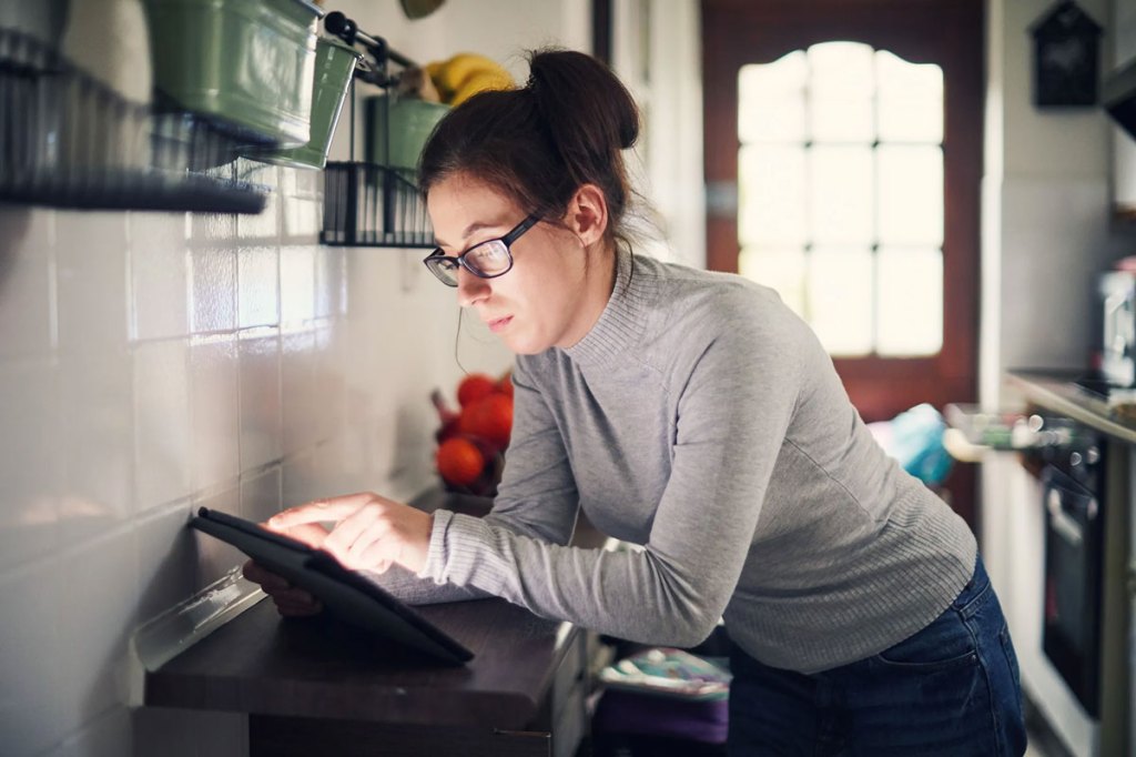 Woman with grey turtleneck and glasses looking at tablet in kitchen