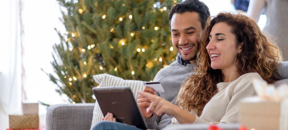 Young couple sitting on couch with a Christmas tree behind them planning their holiday shopping.