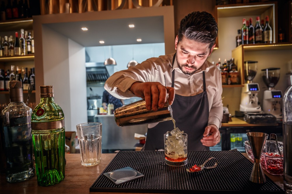 Male bartender is making cocktail pouring alcohol from shaker to glass at bar background