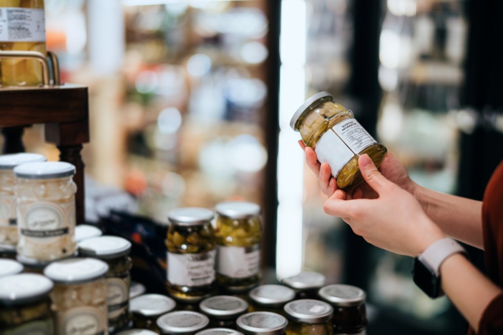 Close-up of a young Asian woman grocery shopping in the supermarket, holding a bottle of preserved vegetables and reading the nutrition label. Making healthier food choices.
