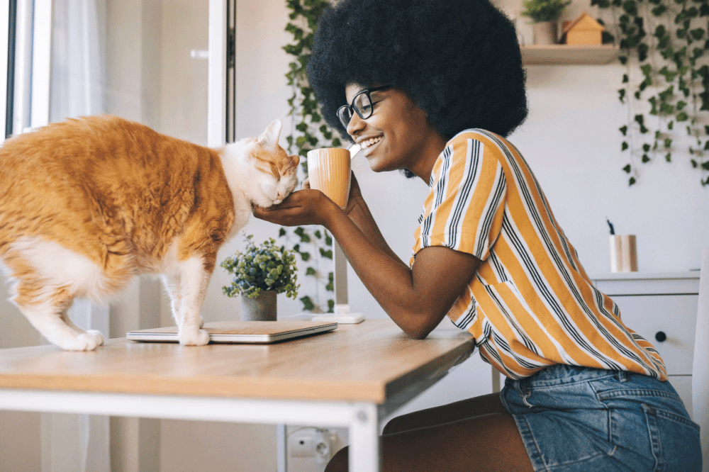 African-American woman with glasses and a striped shirt pets her cat at a table having coffee