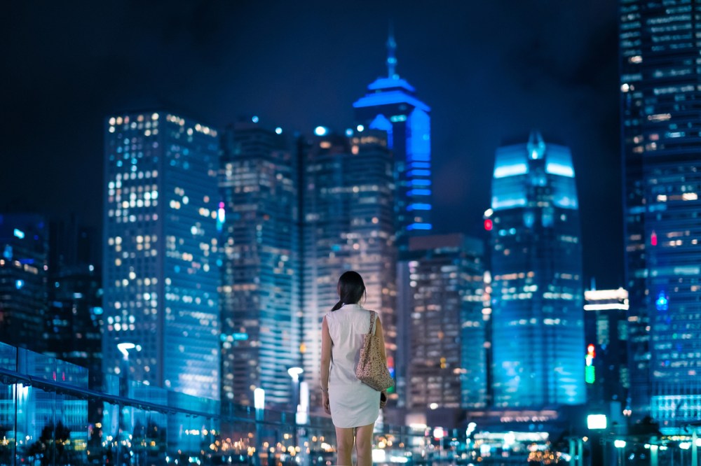 Rear view of Asian female overlooking and enjoying the stunning view of modern city night scene, with massive illuminated corporate highrise buildings in the background.