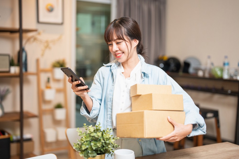 An Asian woman wearing a denim shirt holding her cell phone in one hand and three packages in the other