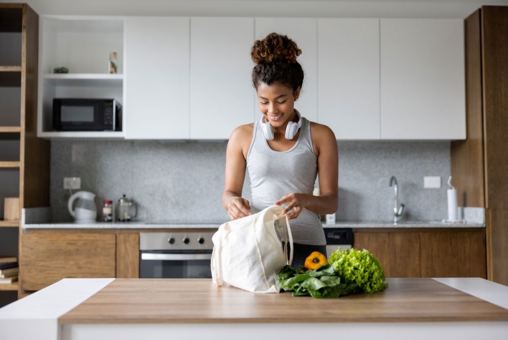 A young woman with athletic ware and headphones unloading groceries on a counter top in a kitchen
