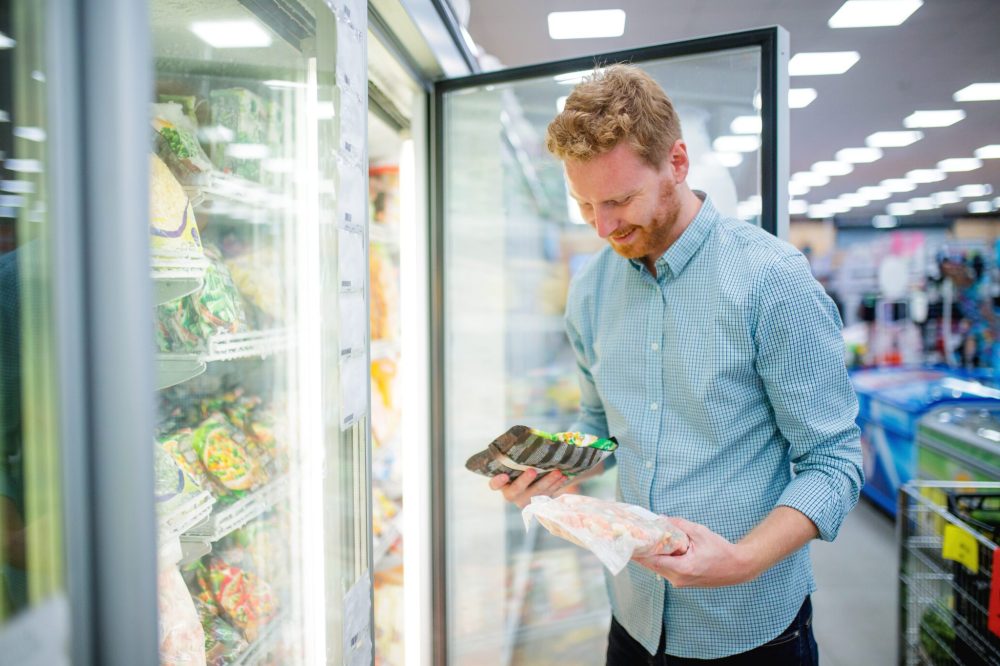 Cheerful young man in supermarket reading nutrion label on frozen food, standing in front of open freezer, copy space