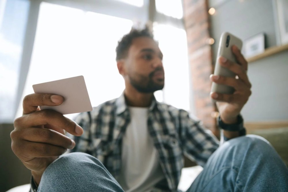 Man in plaid shirt holding a credit card in one hand and his cell phone in another online shopping / ecommerce