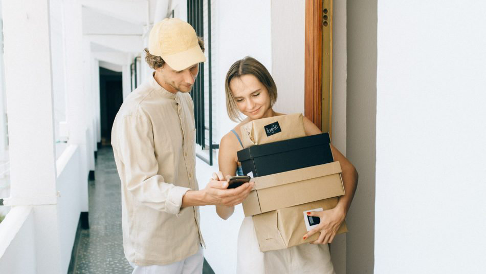 Man with a hat holding mobile phone with woman holding several packages at a doorway