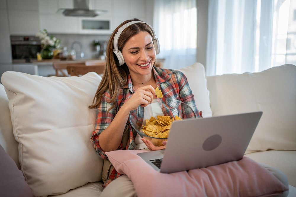The photo shows a young woman sitting comfortably in bed, surrounded by soft pillows, in the living room. A laptop is lying on her lap, and an online movie is being shown on the screen via a TV platform.