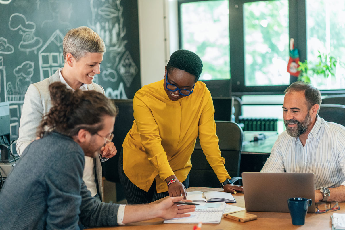 A team works together in an office. The central figure is wearing glasses and has a yellow shirt. There is a blackboard in the background.