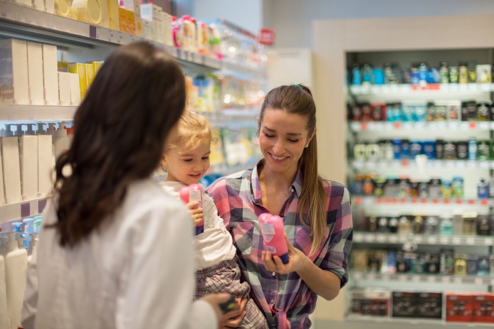 A pharmacist in a white coat talks to a mother who's holding her daughter looking at a product