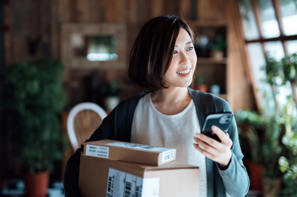 Beautiful smiling young Asian woman with smartphone, receiving parcels with home delivery service at home. Online shopping, mobile payment. Enjoyable shopping experience