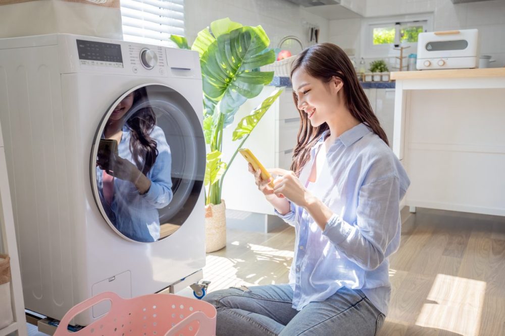 asian woman do the laundry in smart washing machine and use smartphone operate it at home
