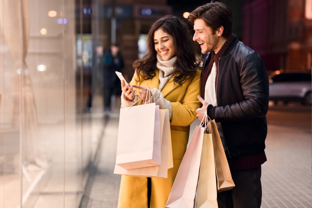 Young couple with smartphone near shopping mall, standing near shop window in the evening;